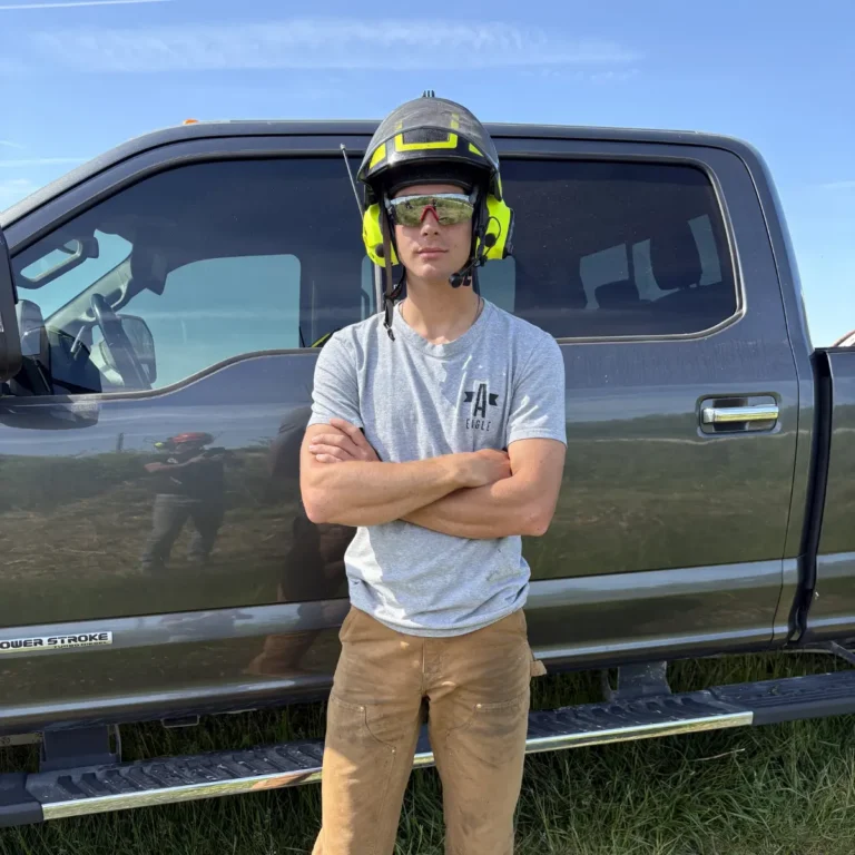 A worker wearing a safety helmet with yellow accent stripes and yellow safety glasses stands with arms crossed in front of a dark-colored crew cab truck under a clear blue sky. The truck bed and grassy ground are visible in the background.