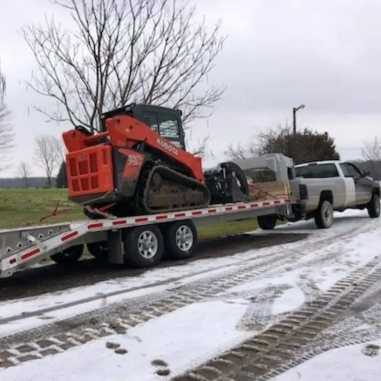 A red skid steer loader on an equipment trailer prepares to load a felled tree log during winter operations. A white pickup truck is parked nearby on a snow-covered lot under overcast skies.