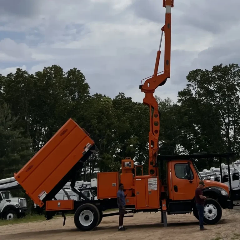 An orange bucket truck with extended boom and a tilted dump truck are positioned in an equipment yard with trees in the background.