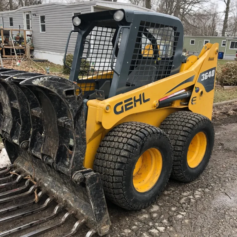 A yellow Gehl skid steer loader with protective cabin cage stands next to a large dark debris pile on a residential work yard with bare trees visible in the background.