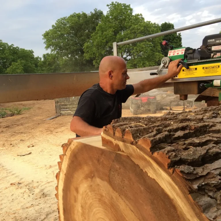 A worker operates a truck-mounted portable sawmill to process logs into lumber sections on a cleared work site. Freshly cut log pieces lay in the foreground with dense trees visible beyond the work area.