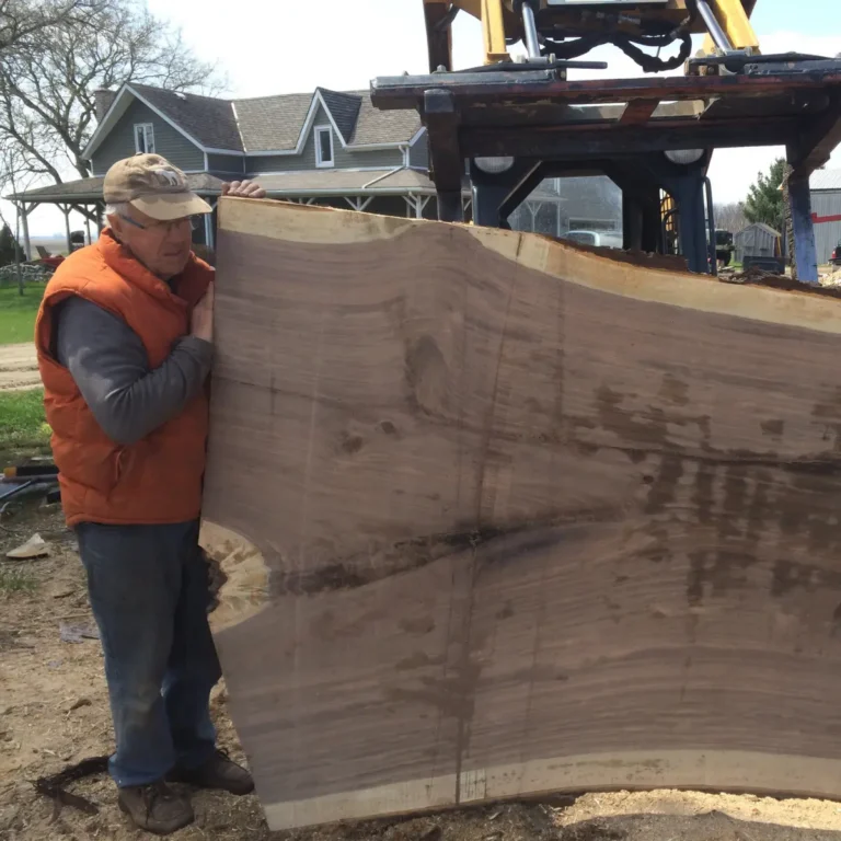 A crew member holds a large milled lumber board at a residential property, with excavator equipment and a pergola structure visible in the background.
