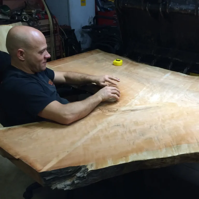 A crew member inspects and measures a large milled wood piece using a tape measure on a workshop workbench.