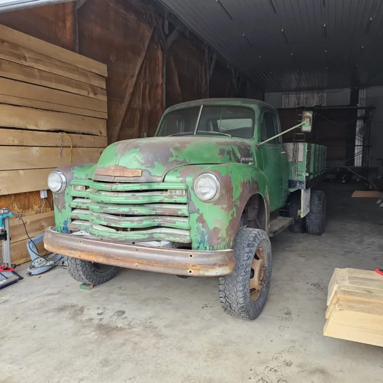 A vintage truck with weathered green paint and rust corrosion parked in a storage barn, equipped with an open stake bed and large work tires. Wooden crates and materials are stacked nearby within the barn.