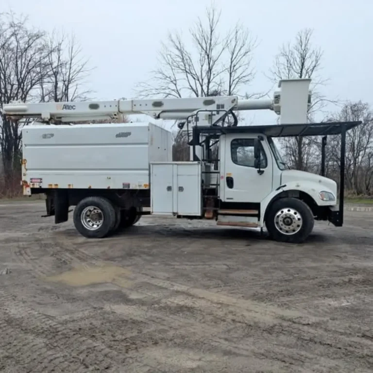 White bucket truck with extended boom parked on an asphalt lot, with bare deciduous trees visible in the background.